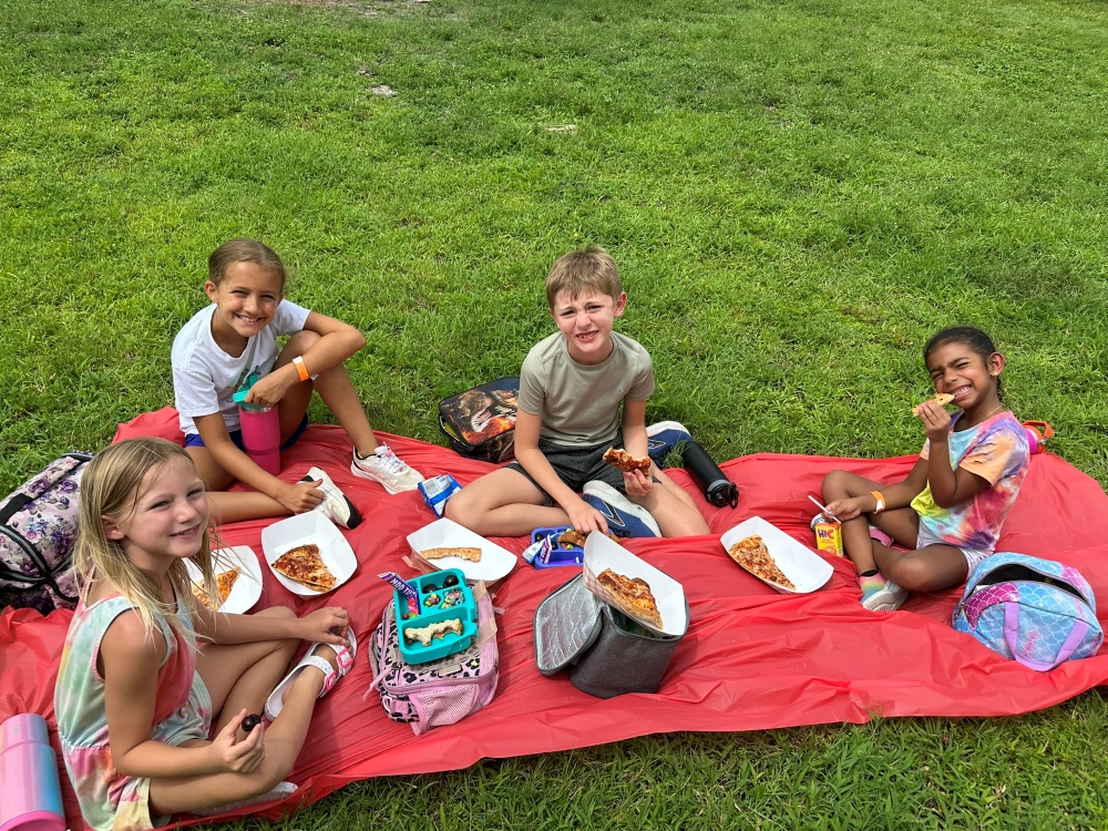 Group of children sit on a red picnic blanket eating pizza on a grassy field